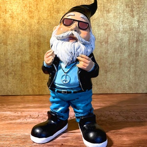 Peut inclure: Un gnome de jardin portant des lunettes de soleil, une veste en cuir, un jean bleu et un collier avec un symbole de paix. Le gnome a une barbe blanche et un chapeau noir.