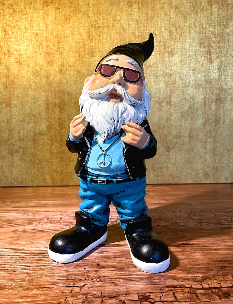 Peut inclure: Un gnome de jardin portant des lunettes de soleil, une veste en cuir, un jean bleu et un collier avec un symbole de paix. Le gnome a une barbe blanche et un chapeau noir.