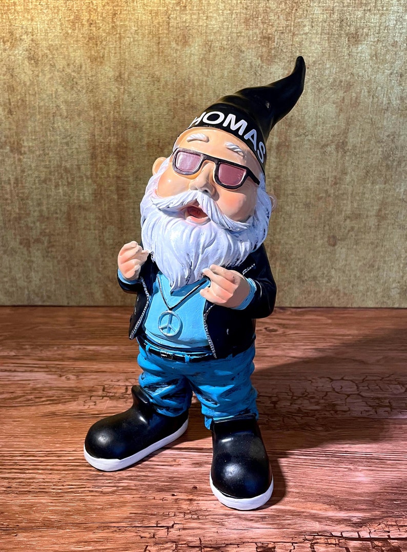 Peut inclure: Un gnome de jardin portant une veste en cuir noire, un jean bleu et des lunettes de soleil. Le gnome a une barbe blanche et un collier avec un symbole de paix. Le chapeau du gnome a le mot "OMAS" &eacute;crit dessus.