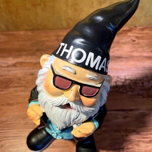 Peut inclure: Un gnome de jardin noir et blanc portant des lunettes de soleil et un chapeau noir avec le mot "THOMAS" dessus. Le gnome porte une veste en cuir noire et un jean bleu.