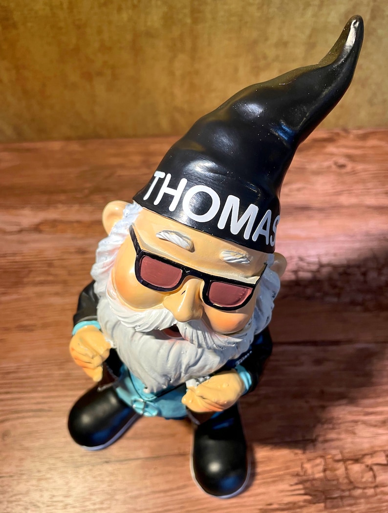 Peut inclure: Un gnome de jardin noir et blanc portant des lunettes de soleil et un chapeau noir avec le mot "THOMAS" dessus. Le gnome porte une veste en cuir noire et un jean bleu.