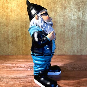 Peut inclure: Une figurine de gnome de jardin bleue et noire portant une veste en cuir, un jean bleu et des baskets noires. Le gnome a une barbe blanche et porte des lunettes &agrave; monture noire.