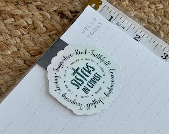 May include: A white sticker with a green design that says "Sisters in Christ" with bible verses around the edge. The sticker is on a white planner with a ruler.