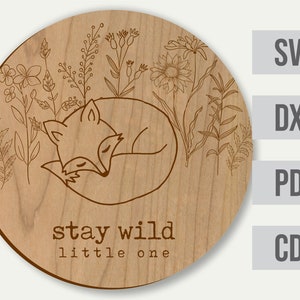 Op de afbeelding: Een rond houten bord met een slapende vos omringd door bloemen en de tekst "stay wild little one".