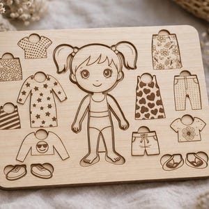 May include: Wooden puzzle showing a girl figure and clothing pieces. The puzzle includes tops, dresses, trousers, shorts, and shoes. The clothing items have patterns like stars, hearts, and floral designs. The puzzle is made of light-coloured wood.
