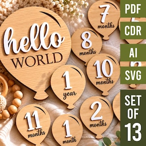 May include: A set of 13 wooden balloon-shaped milestone markers featuring white 3D numbers and text. The largest piece displays "hello WORLD" while others show "1 month" through "1 year". The set includes digital files labeled PDF, CDR, AI, and SVG.