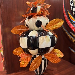 May include: A decorative tabletop ornament featuring a stack of three pumpkins in varying sizes. The pumpkins are painted with black and white checkered and spotted patterns, accented with metallic orange leaves and a black and white striped base.