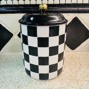 May include: A ceramic jar with a black and white checkered pattern. The jar has a black lid and a gold-colored knob. The jar is sitting on a speckled countertop, with a black and white tile backsplash.