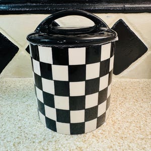 May include: A black and white checkered ceramic jar with a black handle on top. The jar has a lid and is cylindrical in shape. The background features black and white tiles.