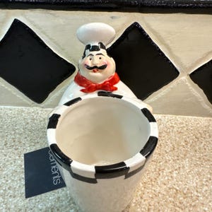 May include: A white ceramic utensil holder shaped like a chef, with a red bow tie and a black and white checkered design. The holder has a wide opening and a small rectangular tag with the word "elements" on it.