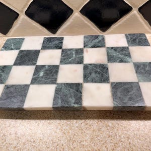 May include: A rectangular serving board featuring a checkerboard pattern made of alternating dark green and white marble tiles. The surface is smooth and polished, showcasing the natural veining and color variations inherent in the stone material.
