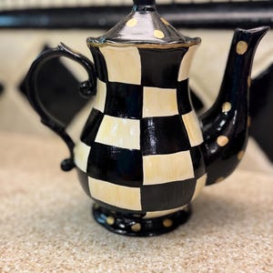 May include: A ceramic teapot featuring a black and cream checkered pattern across the body. The lid, handle, and spout are solid black with cream-colored polka dots. The teapot has a curved handle, a tapered spout, and a matching lid with a finial.