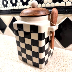 Puede incluir: Un recipiente de cerámica cuadrado con un patrón de cuadros blancos y negros, rematado con una tapa de madera redonda y un pequeño pomo de madera. Una cuchara de madera se sujeta en un asa lateral, asegurada con un cordel al cuerpo.