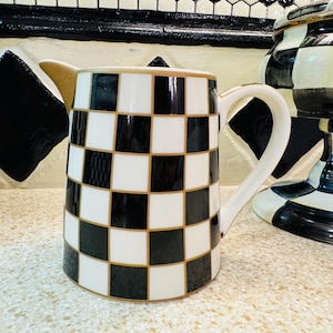 May include: A ceramic pitcher with a black and white checkered pattern. The pitcher has a curved handle and a spout with a gold trim. The design is repeated on other decorative items in the background.