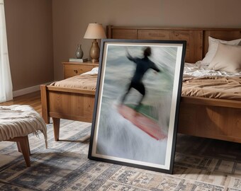 Può includere: Una stampa artistica incorniciata con un surfista su un'onda. Il surfista, in una muta nera, è sfocato, suggerendo movimento. La tavola da surf è rossa. La stampa è in una cornice nera, appoggiata a un letto in una camera da letto.