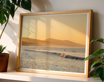 May include: A framed print of a coastal scene at sunset. The image features a wave breaking in the ocean, with several figures surfing. The sky is a warm golden color, and the frame is made of light-colored wood.