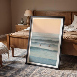May include: A framed art print depicting a seascape with surfers. The artwork has a black frame and shows a calm ocean scene with blue waves and a peach-coloured sky. The print is leaning against a wooden bed frame.