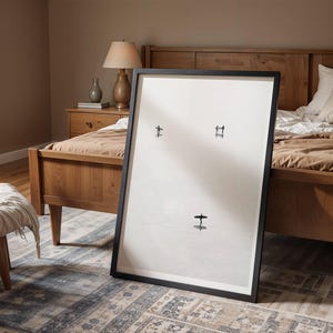 Puede incluir: Una obra de arte grande y enmarcada con un borde negro, apoyada contra una cama. La obra presenta tres pequeñas formas negras abstractas sobre un fondo blanco. El dormitorio tiene una estructura de cama de madera, una mesita de noche y una alfombra estampada.