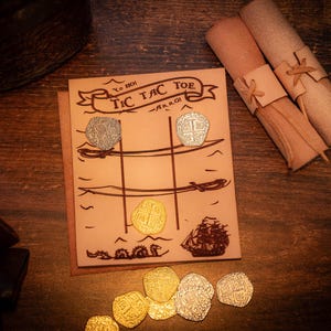 May include: A brown leather game board with the words "Tic Tac Toe - Ahoy!" printed on it. The board has a grid with three rows and three columns. There are gold and silver coins on the board, and a small ship illustration in the bottom right corner.