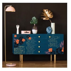Può includere: Una cassettiera blu scuro con maniglie dorate e decalcomanie floreali. La cassettiera ha quattro cassetti e poggia su quattro gambe dorate. La cassettiera è decorata con un motivo floreale in tonalità arancio e nero. La cassettiera è davanti a uno sfondo scuro.