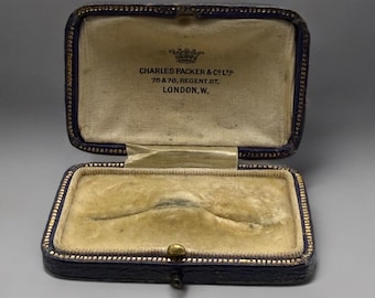 May include: Open, antique jewelry box with a navy blue exterior and gold trim. The interior is lined with a cream-colored velvet. The box is open, revealing the text "CHARLES PACKER & CO LTD 76 & 78, REGENT ST, LONDON, W."