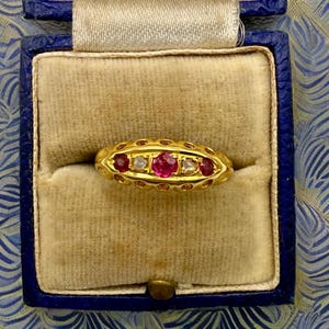 May include: A gold ring featuring a row of five stones, including three round red rubies and two clear white diamonds, set within a decorative yellow gold band with a scalloped edge, presented inside a blue velvet-lined jewelry box.