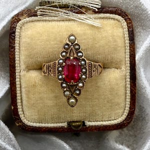 May include: A vintage gold ring with a central, rectangular red gemstone, surrounded by small pearls. The ring is set in an ornate, filigree design and is displayed in a brown velvet ring box. The ring's band features etched details.