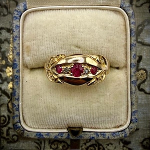 May include: A gold ring featuring a central row of three round red rubies interspersed with two small clear stones. The band has a polished finish with decorative, textured metal detailing on both sides of the central stone setting.