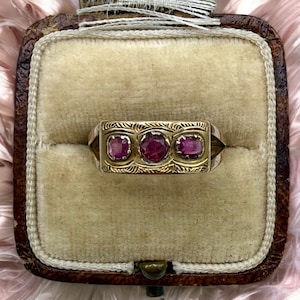May include: A gold ring featuring three round, deep pinkish-red rubies set in a horizontal row. The metal band displays intricate, engraved leaf-like patterns surrounding each gemstone, creating a textured surface across the front of the ring.
