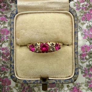 May include: A gold ring featuring a central round-cut vibrant pink stone flanked by two smaller round-cut pink stones, with two tiny clear stones set between them, all arranged in a horizontal row within a cream-colored velvet jewelry box.