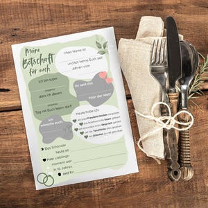 Puede incluir: Un juego de papelería con temática de boda con un diseño decorativo, que presenta una tarjeta para rellenar y una servilleta con cubiertos atados con hilo. La tarjeta tiene texto en alemán. Los artículos están dispuestos sobre una superficie de madera.