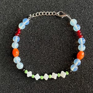 May include: A beaded bracelet featuring a mix of colorful beads. The bracelet includes iridescent blue, red, orange, white, and green beads, connected by a silver chain and clasp. The bracelet is displayed on a black surface.