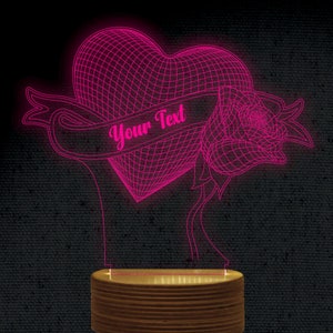 Puede incluir: Un corazón rosa impreso en 3D con una rosa y una pancarta que dice "Your Text". El corazón, la rosa y la pancarta están iluminados con luz rosa. La base es de madera.