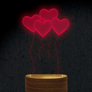 Puede incluir: Lámpara decorativa con cuatro corazones rojos de alambre que flotan sobre una base de madera. Los corazones están iluminados con un brillo rojo brillante, creando una estética romántica. La base tiene un acabado de madera natural y cálido.
