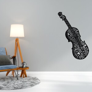 May include: A black and white patterned cello wall decor with intricate details. The cello is mounted on a white wall with a blue armchair and a lamp in the foreground.