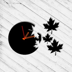 Puede incluir: Reloj negro con manecillas naranjas y un diseño de hojas de arce recortadas en la esfera. El reloj está montado sobre un fondo de madera blanca.