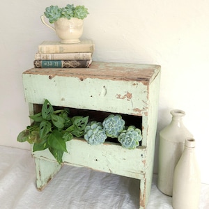 May include: A light green wooden stool with a distressed finish and an open storage compartment. The stool holds a stack of three books, a cream-coloured teacup with a succulent, and a green plant. Two cream-coloured ceramic bottles stand nearby.