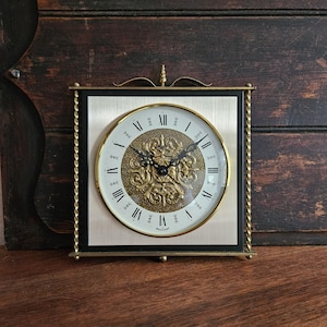 May include: A square wall clock featuring a silver-toned face with black Roman numerals and a central gold-toned embossed floral medallion. The clock is framed by twisted brass-coloured metal pillars and topped with a decorative finial.