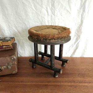 May include: A small, round stool featuring a dark brown wooden frame with four cylindrical legs connected by a cross-shaped base. The seat is circular, covered in a textured, light brown woven material with a frayed, fibrous outer edge.