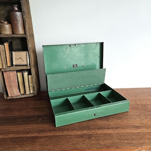May include: A rectangular green metal cash box with an open hinged lid, featuring a central interior divider and four separate compartments for currency storage. The front of the box includes a small keyhole for locking the metal container securely.