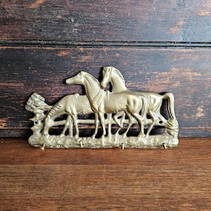 May include: A brass key holder depicting three horses. One horse is grazing, while the other two are standing. The key holder has four hooks and is mounted on a wooden surface.