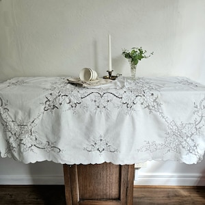 May include: A white rectangular tablecloth featuring intricate black thread embroidery of grapevines and leaves along the edges and centre. The scalloped border frames the fabric, which is draped over a wooden surface with a candle and vase.