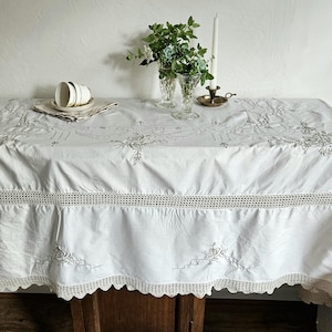 May include: A white rectangular tablecloth featuring intricate grey floral embroidery, a horizontal band of openwork crochet, and a scalloped crochet border. Two white ceramic cups sit on a matching napkin, with two glass vases and a candle.