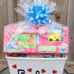 Puede incluir: Una cesta de regalo blanca con un lazo azul en la parte superior. La cesta est&aacute; llena de juguetes de Cocomelon, incluyendo una alfombra de juego, un autob&uacute;s y un peluche. La cesta est&aacute; etiquetada con "Easton".
