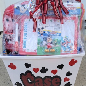 Puede incluir: Una cesta de pl&aacute;stico blanca con dise&ntilde;os de Mickey Mouse rojos y negros. La cesta est&aacute; llena de una variedad de art&iacute;culos con tem&aacute;tica de Mickey Mouse, incluyendo un juguete de peluche Mickey Mouse rojo y blanco, un libro para colorear Mickey Mouse rojo y blanco, una pizarra blanca de borrado en seco y una figura de Mickey Mouse. La cesta est&aacute; envuelta en pl&aacute;stico transparente con una cinta roja y una cinta de lunares blancos y rojos.