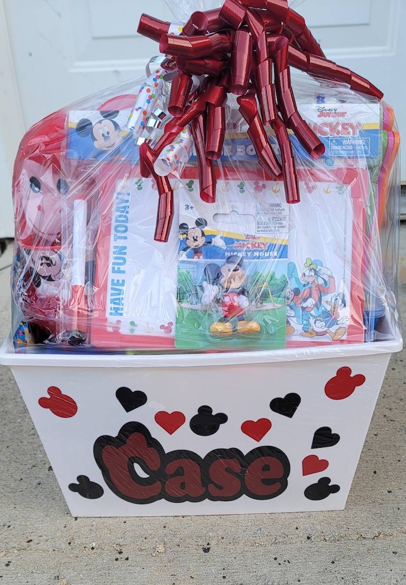 Puede incluir: Una cesta de pl&aacute;stico blanca con dise&ntilde;os de Mickey Mouse rojos y negros. La cesta est&aacute; llena de una variedad de art&iacute;culos con tem&aacute;tica de Mickey Mouse, incluyendo un juguete de peluche Mickey Mouse rojo y blanco, un libro para colorear Mickey Mouse rojo y blanco, una pizarra blanca de borrado en seco y una figura de Mickey Mouse. La cesta est&aacute; envuelta en pl&aacute;stico transparente con una cinta roja y una cinta de lunares blancos y rojos.