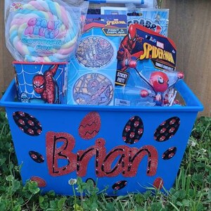 Puede incluir: Una cesta de pl&aacute;stico azul con tem&aacute;tica de Spiderman. La cesta tiene el nombre "Brian" escrito en rojo y negro en la parte delantera. La cesta est&aacute; llena de varios art&iacute;culos con tem&aacute;tica de Spiderman, incluyendo una pegatina de Spiderman, una impresi&oacute;n y una pantalla de Spiderman, un juguete de Spiderman con pies pegajosos y un algod&oacute;n de az&uacute;car de Spiderman.