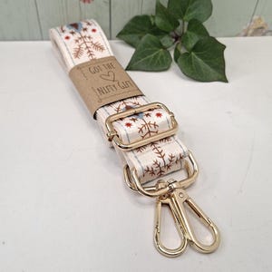 May include: Cream-coloured bag strap with a repeating pattern of red and blue trees and stars. The strap has gold-coloured metal hardware, including a buckle and two swivel clasps. The text "Got the Nifty Gift" is printed on a brown paper band.