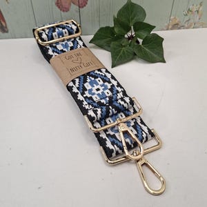 May include: A patterned bag strap in black and white with blue detailing and gold-coloured metal fixings. The strap has a geometric design and a brown paper tag with the text "GOT THE NIFTY GIFT".