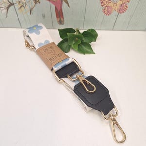 May include: A cream-coloured bag strap with a blue floral pattern and gold-coloured hardware. The strap has a black leather accent and a tag that reads "Got the Nifty Gift".